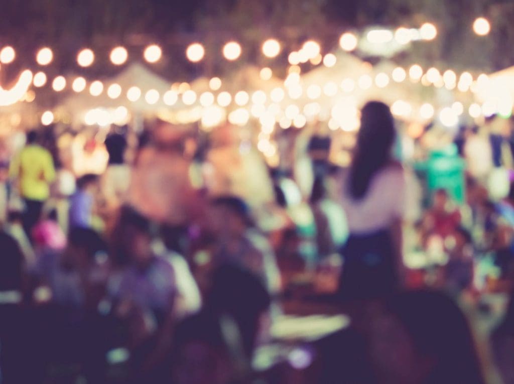 Blurred image of a crowded outdoor event with string lights and a silhouette of a person in the foreground.